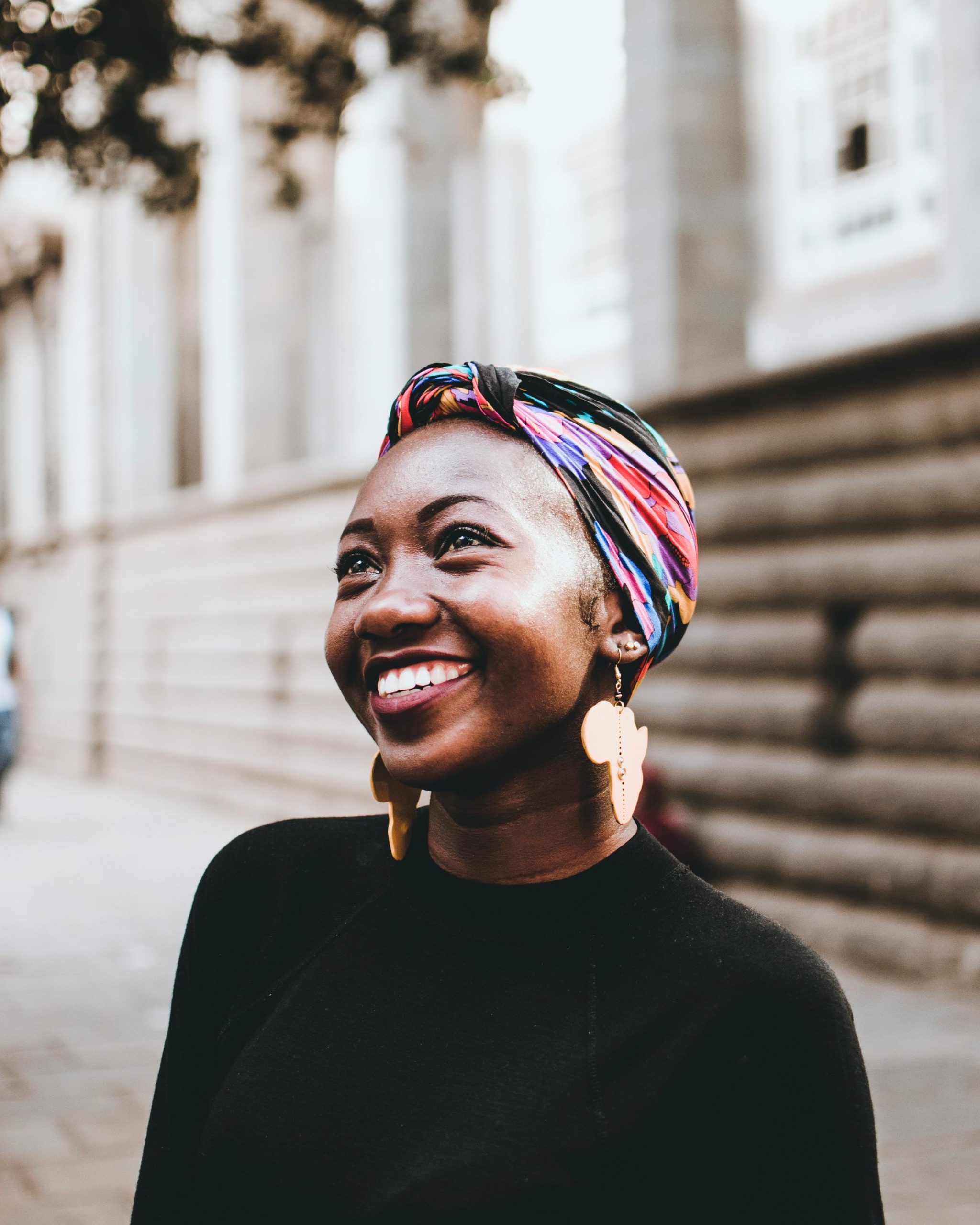 Smiling woman wearing vibrant headscarf and earrings, captured in an urban street setting.